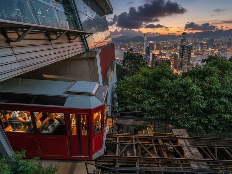 A red Peak Tram carriage at the top station, with the lit towers of Hong Kong and the harbour beyond the trees at dusk